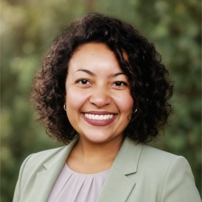 Luana sorrindo, de cabelos cacheados e blazer verde-claro, diante de folhagens.